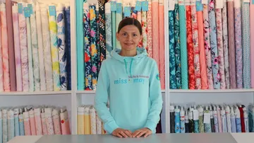 Woman stands in front of colorful rolls of fabric in a shop.