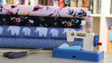 Sewing machine sews fabrics in front of colorful rolls of fabric on the table.
