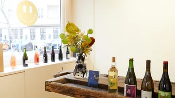 Flower vase stands on table, surrounded by wines and windows.