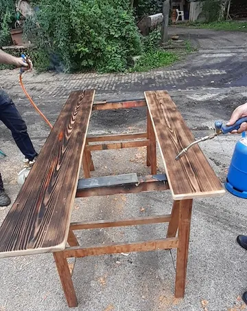 Holztisch wird mit einer Brennerflamme behandelt, draußen.