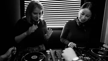 Two DJanes mix music on a DJ stand, illuminated.