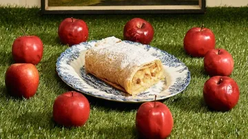 Apfelstrudel liegt auf Teller, umgeben von roten Äpfeln.
