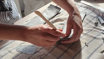 Hände formen Ton mit einem Holzwerkzeug auf Tisch.