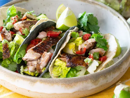Three tacos with chicken, vegetables and lime slices in a bowl.
