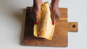 Hände, die Kochbananen auf einem Holzbrett schälen.