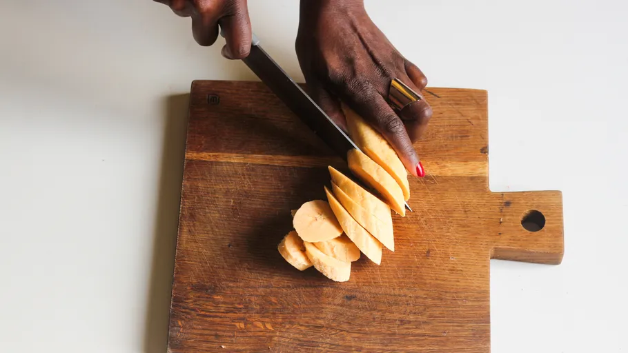 Hand slicing sweet potatoes on wooden board.