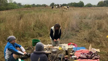 Three people sit on the ground while one cooks. Cows graze in the distance.