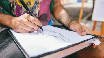 Eine Person zeichnet mit einem schwarzen Stift auf einem großen Skizzenblock, der auf ihrem Schoß liegt, konzentriert auf die Illustration.