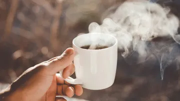 One hand holds a steaming cup of coffee outside.