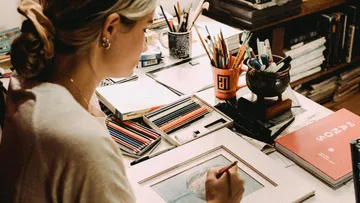 Woman draws on a table full of drawing materials.