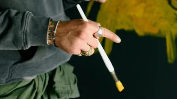 Ein Hand hält einen Pinsel über eine gelbe Fläche.