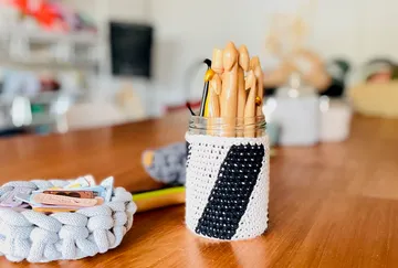 Glas mit Häkelüberzug hält kreative Werkzeuge auf Tisch.