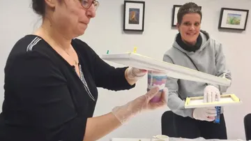 Two women hold art materials in a bright room.