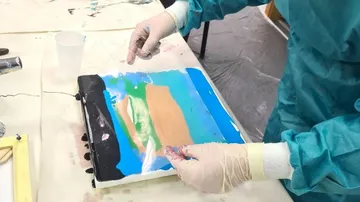 Hands place paint on a transparent sheet in a room.