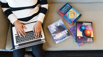 Eine Person tippt auf einem Laptop, umgeben von Lehrbüchern.