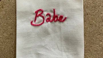 Gesticktes Wort „Babe“ auf weißem Stoff, helles Umfeld.