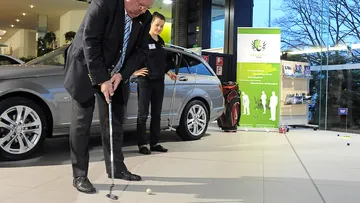 Mann spielt Golf im Autohaus, während Zuschauer zuschauen.