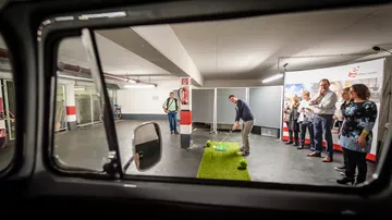 Golfer schlägt auf Kunstrasen in Parkgarage; Zuschauer beobachten.