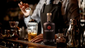Rumflasche steht auf der Bar, Barkeeper mixt Getränke im Hintergrund.