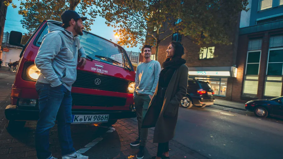 Rotes VW-Bus, drei Personen unter Bäumen, Abendstimmung.