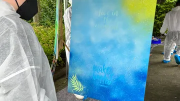Eine Person sprayt auf eine farbige Leinwand im Freien.