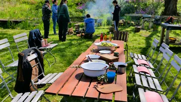 Ein gedeckter Tisch mit Schüsseln, Menschen bereiten Essen im Garten.