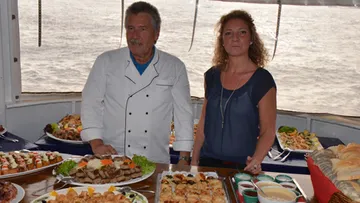 Koch und Frau stehen neben Buffet auf einem Boot.