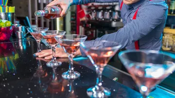 Barkeeper füllt rosa Cocktails in Martini-Gläser auf einer glänzenden Theke, im Hintergrund bunte Barutensilien.