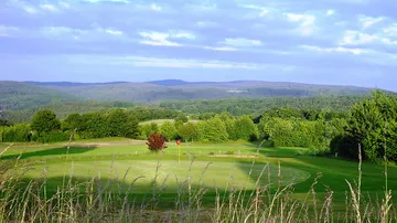 Green golf course fairway with flag against a hilly forest landscape.