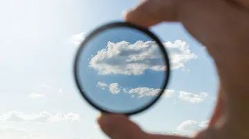 Object: A hand with a filter. Action: The filter shows clouds in the sky. Context: Bright blue sky with clouds.