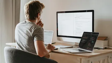 Mann sitzt am Schreibtisch, arbeitet an mehreren Computern.