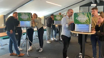 Menschen stehen an Tischen mit Kästen, diskutieren aktiv.