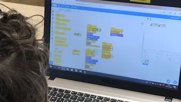A laptop shows programming blocks. One person is working on it.