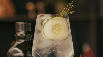 Ein Glas mit Getränk und Gurke, im Hintergrund verschwommene Formen.