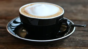 Black cup with cappuccino on saucer on wooden background.