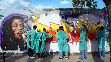 Group of people in green coats spray graffiti on a wall showing 'Berlin' and a portrait of Bob Marley.