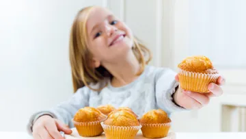 Girl holding muffin and smiling, other muffins next to it.