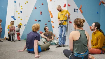 Climber stands with equipment, group sits and listens.