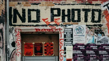 Graffiti an einer Wand zeigt `NO PHOTO`, umgeben von Plakaten.