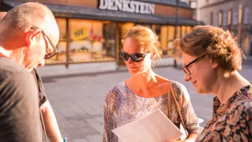 Three people are looking at something in a town square in sunlight.
