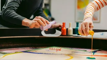 Paints are poured onto a painting surface by two people.