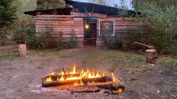 Lagerfeuer brennt vor einem Holzhaus in der Natur.