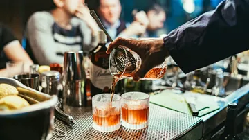 Ein Barkeeper gießt Cocktails in Gläser. Menschen im Hintergrund.
