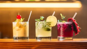 Drei Cocktails stehen auf einer Holztheke, im Hintergrund Beleuchtung.