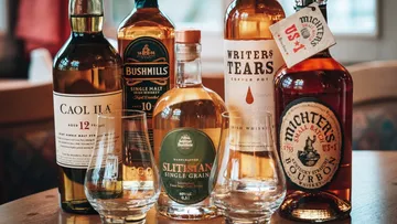 Sechs Whiskeyflaschen stehen zusammen mit Gläsern auf einem Tisch.