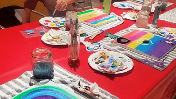 Farbenfrohe Malutensilien liegen auf einem roten Tisch.