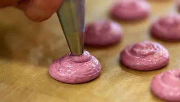 Eine Spritztüte formt rosa Makronen auf einer Unterlage.