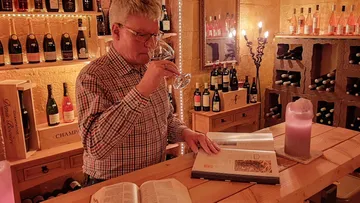 Mann riecht Wein und liest in einem Buch in einem Weinkeller.