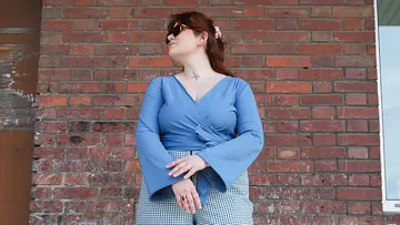 Frau in blauer Bluse steht vor Ziegelwand, schaut nach oben.