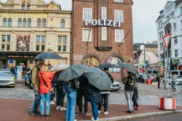 Gruppierung von Menschen mit Regenschirmen vor Polizeigebäude.